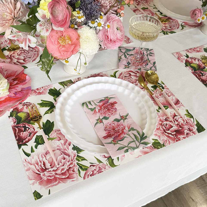 A table set with Hester & Cook Peonies in Bloom Placemats, white plates, gold cutlery, a glass of champagne, assorted flowers as a centerpiece, and a white tablecloth.