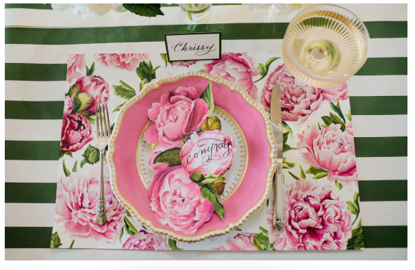 A place setting with a pink floral plate and Hester & Cook Peonies in Bloom Placemat, gold-rimmed glass, silverware, and a name card reading “Chrissy” on a green and white striped tablecloth.