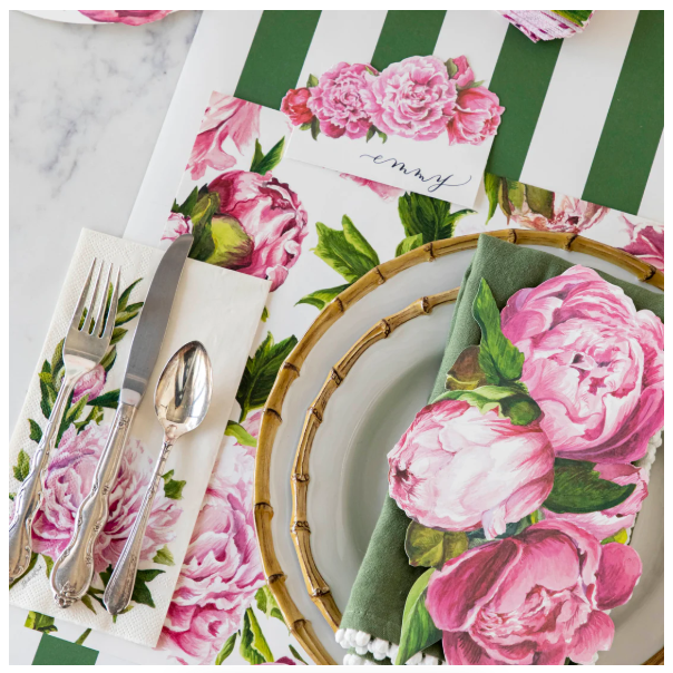 A table setting with a pink peony plate, silverware, and napkin rests on Hester & Cook’s Peonies in Bloom Placemat (Pad of 18), finished with a floral place card reading “emmy.”.