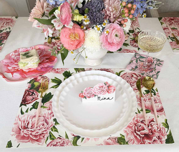 A floral-themed table setting features a white plate, gold cutlery, wine glass, and name card for Nina. The napkin and centerpiece of assorted flowers are adorned with Hester & Cook’s Peonies in Bloom Placemat - Pad of 18 Sheets.