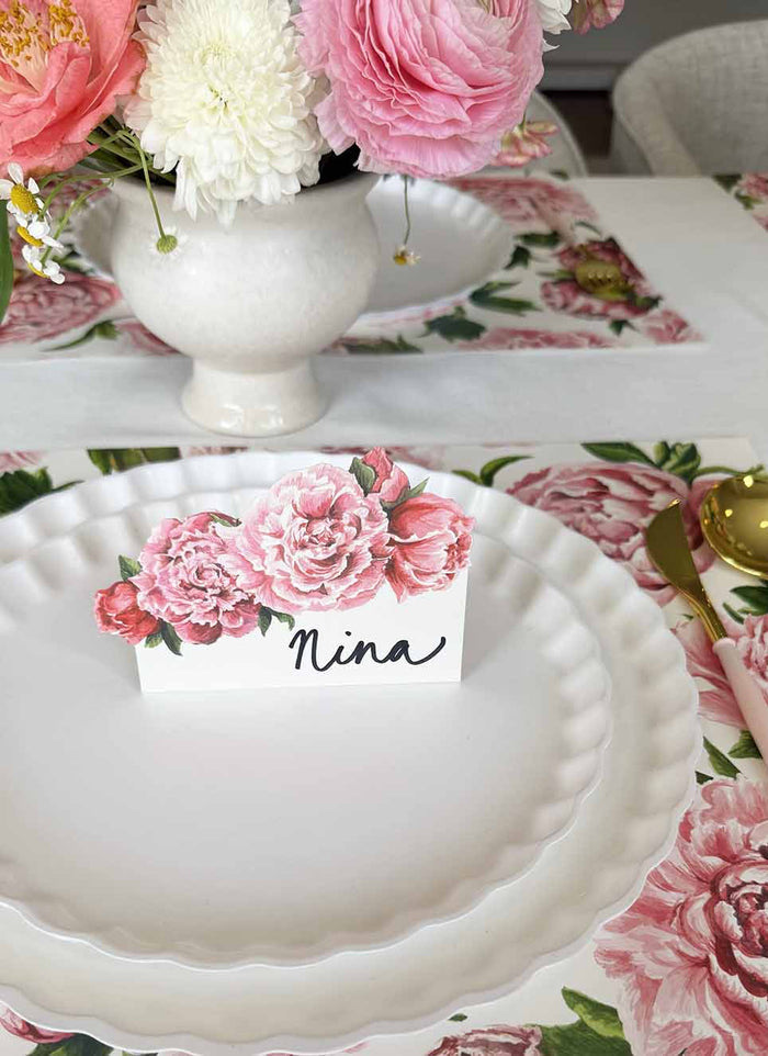A scalloped white plate with a Hester & Cook Peony Place Card—set of 12—reading “Nina” sits on a pink peony placemat. A white vase of flowers and gold Hester & Cook cutlery complete the elegant table setting.