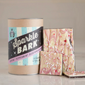 A canister labeled Peppermint Sparkle Bark by Hester & Cook with white and pink peppermint chocolate swirled bark, topped with crushed candy canes on a light surface. Gluten free.