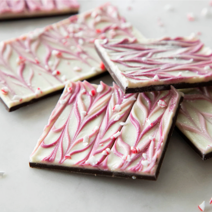 Peppermint Sparkle Bark by Hester & Cook features gluten-free chocolate squares with a dark Belgian chocolate base, white chocolate top swirled in pink and white, and candy bits, artfully displayed on a light surface.