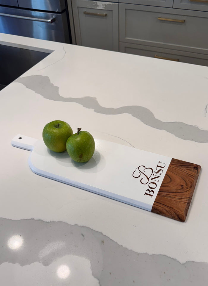 A Maple Personalized Acacia Bevel Board in white (20" x 7" x .6") sits on a marble kitchen counter with "BONSU" engraved and two green apples. Kitchen cabinets and an oven are visible in the background.