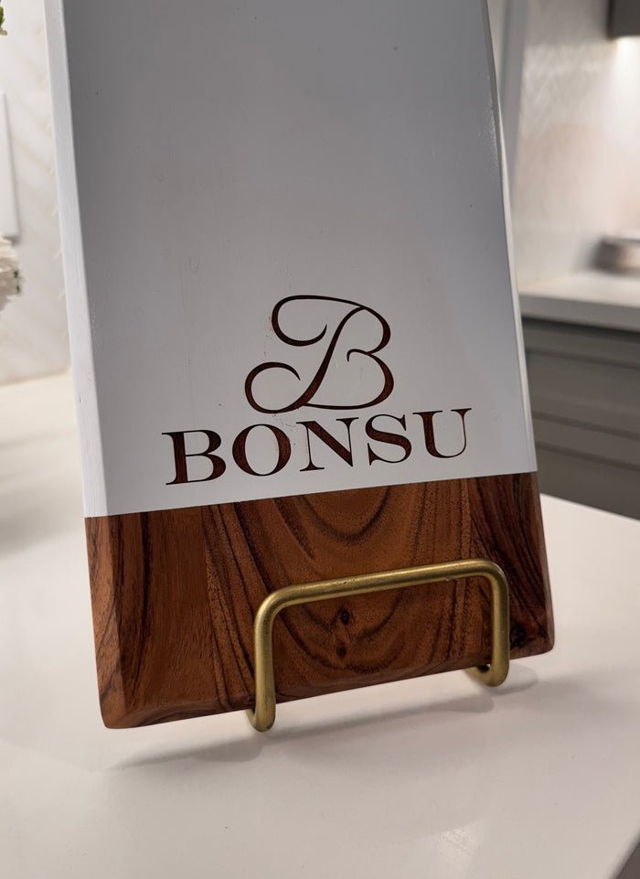 The Maple Personalized Acacia Bevel Board (White, 20" x 7" x .6") with “BONSU” and a decorative “B” stands on a brass stand in a modern kitchen.