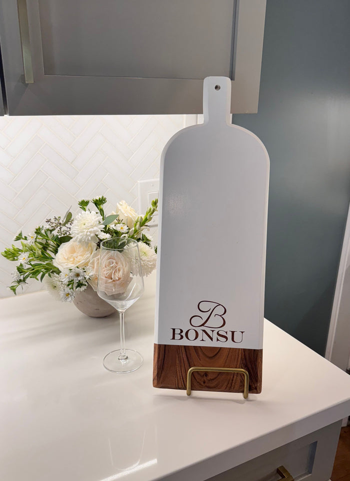 A white kitchen counter displays the Maple Personalized Acacia Bevel Board (20" x 7" x .6", White) upright on a stand, alongside a wine glass and a vase of white flowers in the background.
