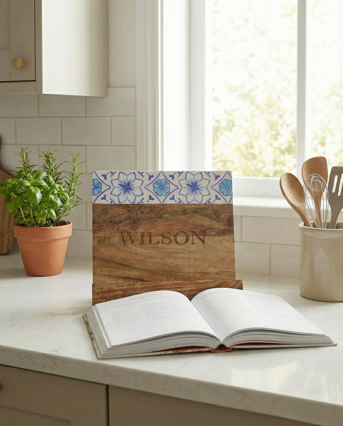 The Maple Personalized Acacia Cookbook Holder, featuring Capri pattern enamel (11x11"), displays an open book on a kitchen counter near a potted herb, utensils, and a sunlit window.