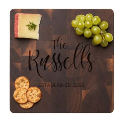 The Maple Personalized Black Walnut Butcher Block Slim Cutting Board (12x12") features "The Russells Established 2022" in script and is pictured topped with a cheese wedge, green grapes, and three crackers.