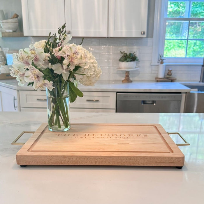 A glass vase with white flowers rests on the Maple Personalized Maple Wood Cutting + Cheeseboard (Test), set on a white kitchen island in a kitchen with white cabinetry and a window overlooking greenery.