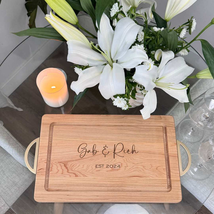 A Personalized Maple Wood Cutting + Cheeseboard (Test) by Maple, engraved with Gab & Rich EST 2024, rests on a glass table beside white lilies, a lit candle, and empty wine glasses.