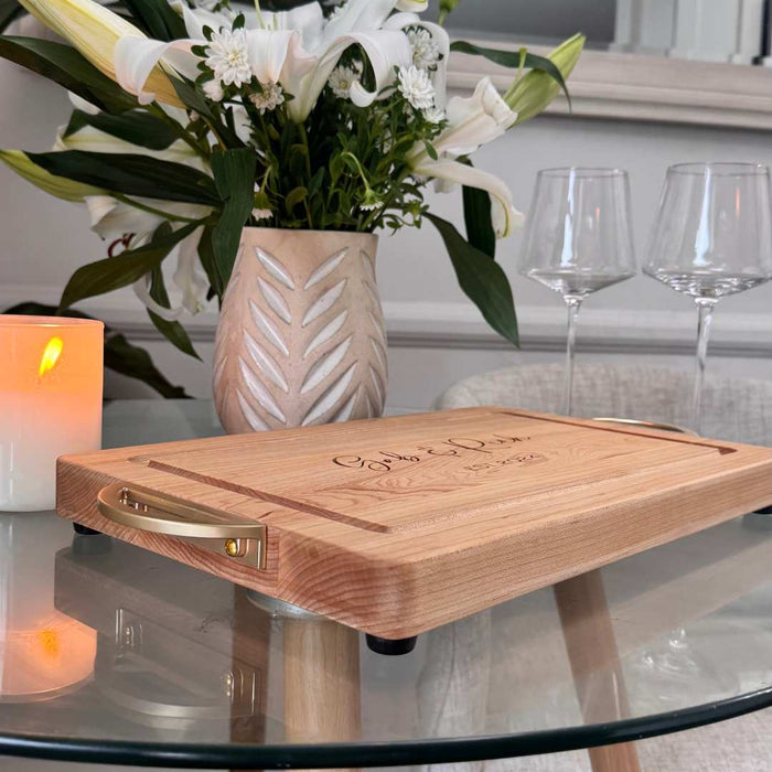 The Maple Personalized Maple Wood Cutting + Cheeseboard (Test) with brass handles rests on a glass table near a lit candle, two empty wine glasses, and a vase of white lilies in a decorative pot.