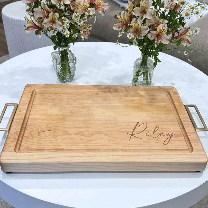 The Maple Personalized Maple Wood Cutting + Cheeseboard 18 x 12" features “Riley” engraved in cursive on a corner, metal handles, and sits on a white table with two flower vases in the background. Perfect for serving charcuterie.