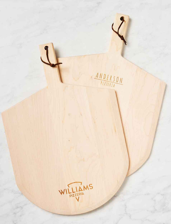 Two Personalized Maple Wood Pizza Peels 14 x 21" by Maple, each with a leather hanging strap and custom names—ANDERSON PIZZERIA and WILLIAMS PIZZERIA—displayed on a white marble surface.