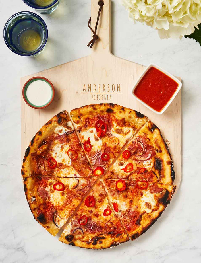 A sliced pepperoni pizza sits on the Maple Personalized Maple Wood Pizza Peel 14 x 21", with two dipping sauces and glasses nearby on a white marble surface.