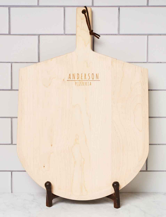 The Personalized Maple Wood Pizza Peel 14 x 21" by Maple stands upright on a metal holder against a white subway tile wall, featuring custom engraving.