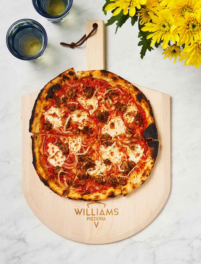 A thin-crust pizza with cheese, onions, and sausage rests on a Maple Personalized Maple Wood Pizza Peel 14 x 21", engraved with Williams Pizzeria. Two glasses of water and a bouquet of yellow flowers are nearby.