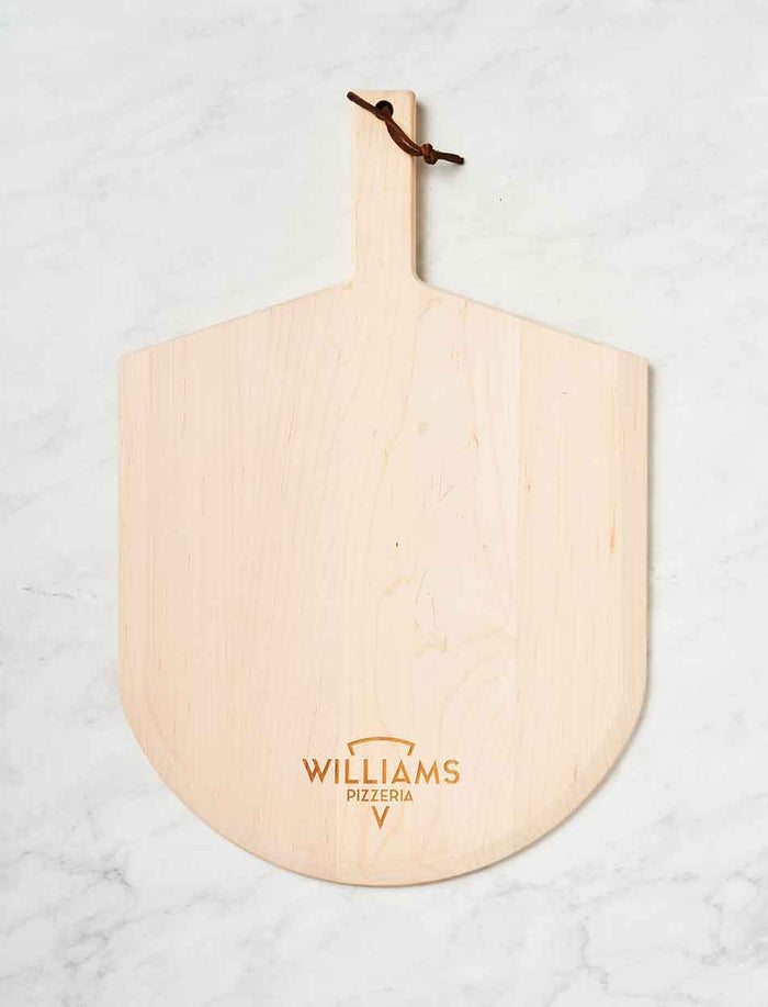The Personalized Maple Wood Pizza Peel 14 x 21" by Maple, featuring a handle and leather loop, displays the brand logo near the bottom and rests on a white marble surface.