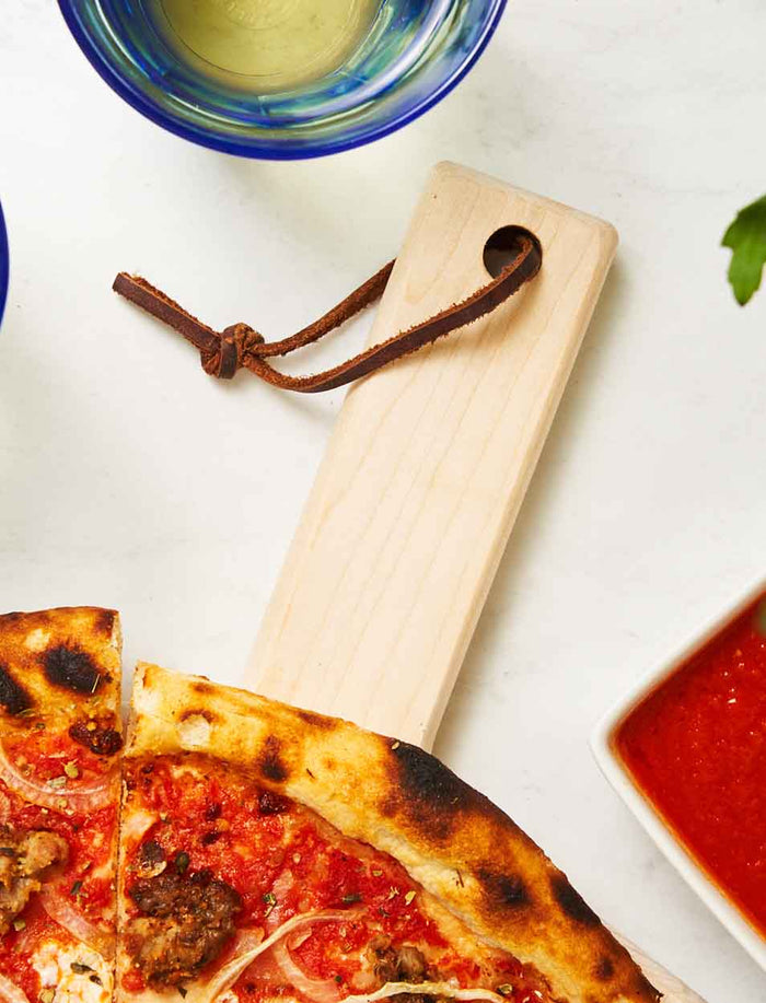 The Maple Personalized Maple Wood Pizza Peel 14 x 21" with a leather strap holds a pizza with crispy crust; a cup of white wine and a square dish of tomato sauce sit nearby on a white surface.
