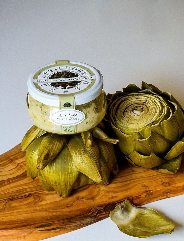 A jar from Bella Cucina's Piccolo Pesto Gift Collection rests on a whole artichoke beside a halved one on a wooden board, with a loose petal in the foreground—showcasing one of their best-loved pesto flavors.