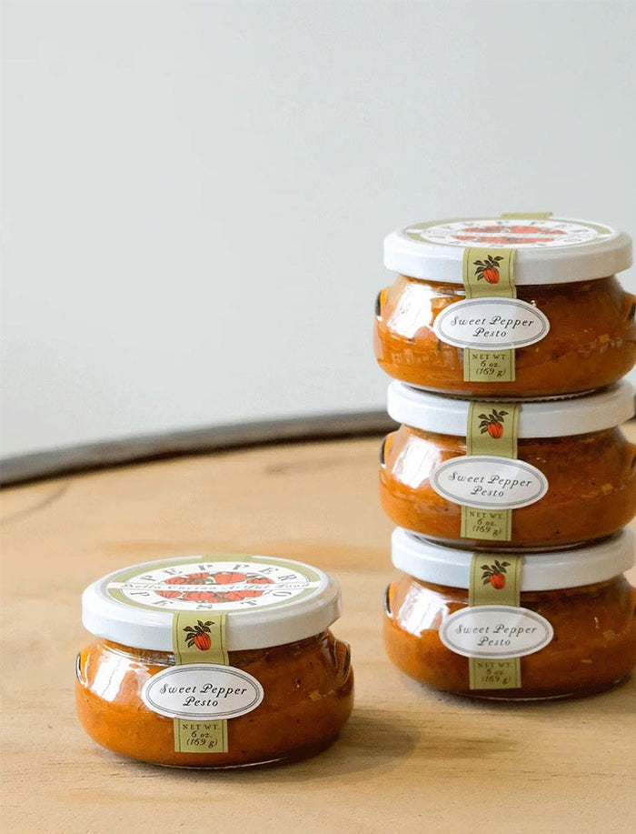 Four small jars from the Bella Cucina Piccolo Pesto Gift Collection are stacked on a light wooden surface, featuring popular pesto flavors—ideal for adding to a gift box.