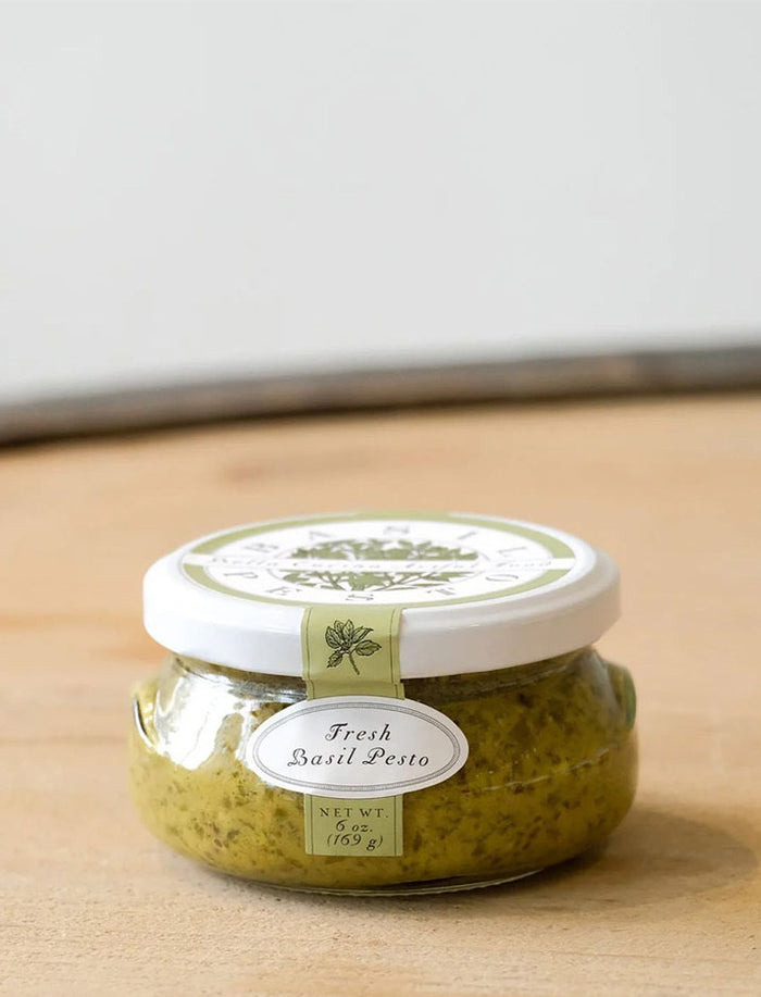 A small glass jar with a white lid, labeled "Fresh Basil Pesto, Net Wt. 6 oz (169 g)," from the Bella Cucina Piccolo Pesto Gift Collection sits on a wooden surface, showcasing one of their popular pesto flavors.