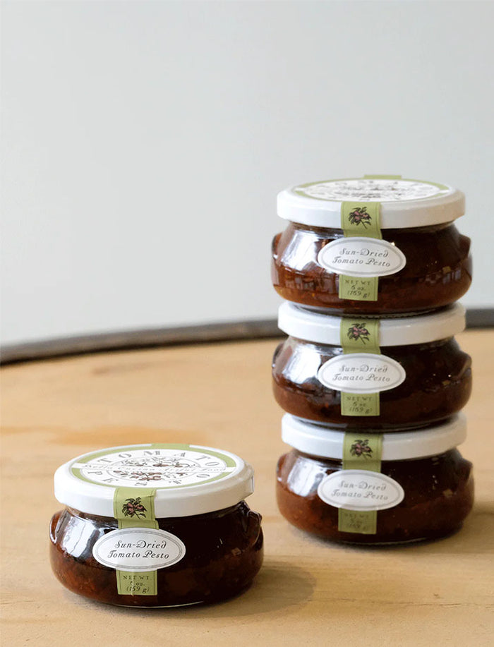 Five jars from the Bella Cucina Piccolo Pesto Gift Collection are displayed on a wooden surface, with four stacked on the right and one set apart on the left. Each jar features a white lid and green label, all against a plain background.