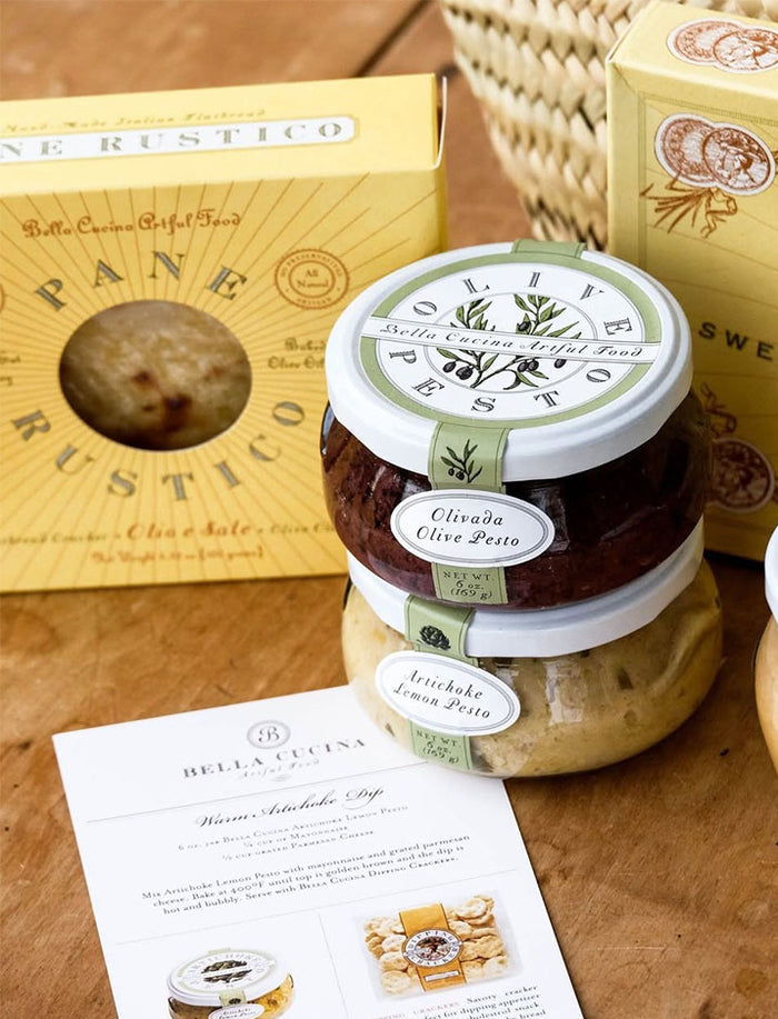 A close-up of the Bella Cucina Piccolo Pesto Gift Collection shows jars of olive pesto and artichoke lemon pesto, a yellow box of rustic Italian bread, and a product information card on a wooden surface.