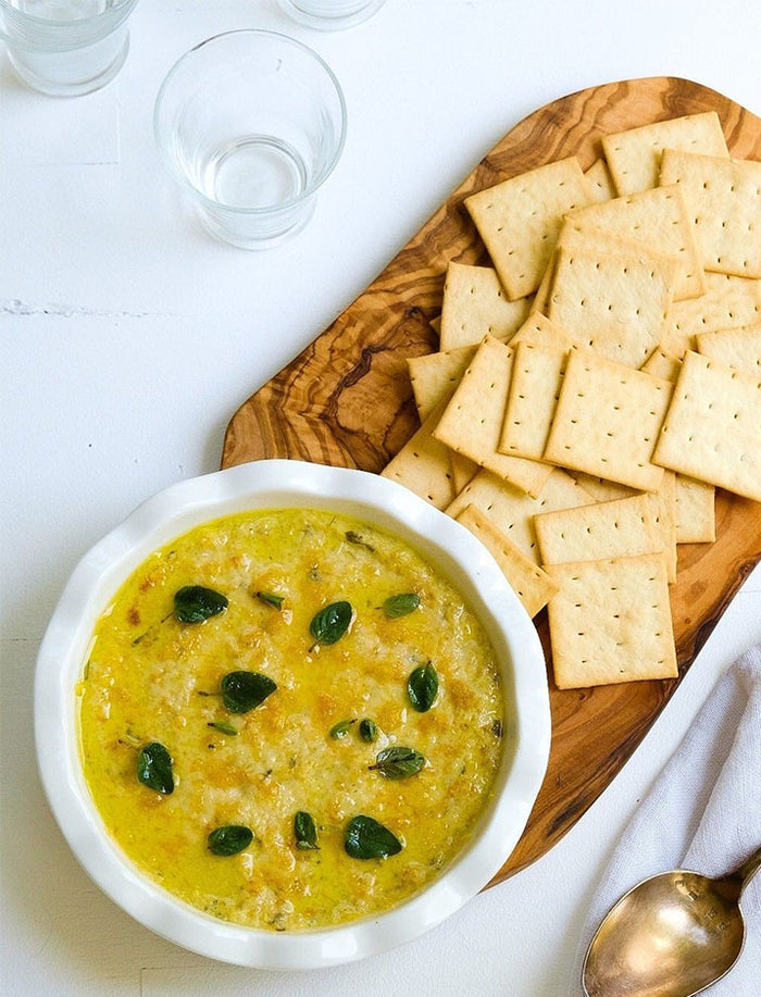 A white dish of cheesy dip garnished with herbs sits beside crackers and a jar from the Bella Cucina Piccolo Pesto Gift Collection, while a spoon and glasses rest on the white surface.