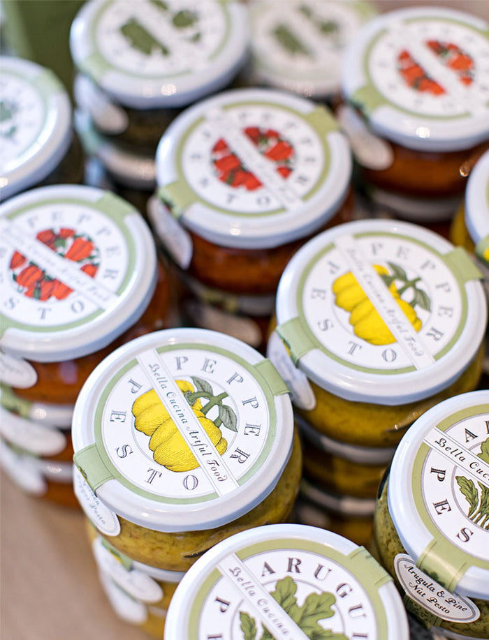 Bella Cucina's Piccolo Pesto Gift Collection features clear glass jars with vibrant vegetable labels like yellow peppers and tomatoes, neatly stacked to showcase the colorful contents inside.
