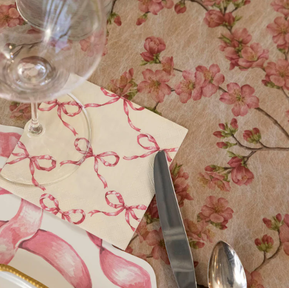 A close-up of a table setting with a floral tablecloth, wine glass, cutlery, and a pink plate features Hester & Cook's Pink Bow Lattice Cocktail Napkins (Pack of 20), adding charm with their delicate pink ribbon pattern.