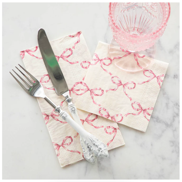 A fork and knife with ornate white handles rest on a Hester & Cook Pink Bow Lattice Guest Napkin (Pack of 16), beside a matching napkin and pink-tinted glass atop a white marble surface.
