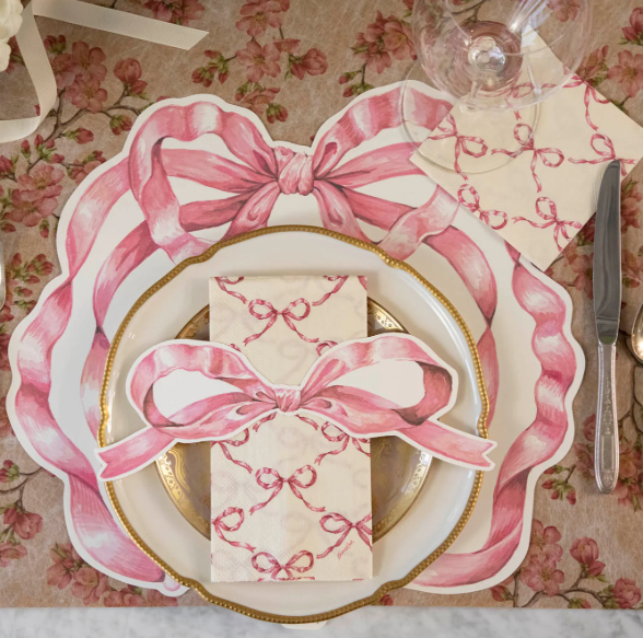 A table setting with a floral tablecloth features Hester & Cook's Pink Bow Lattice Guest Napkins (Pack of 16), gold-rimmed plates, wine glass, fork, and knife for a feminine, elegant style—perfect for guests.