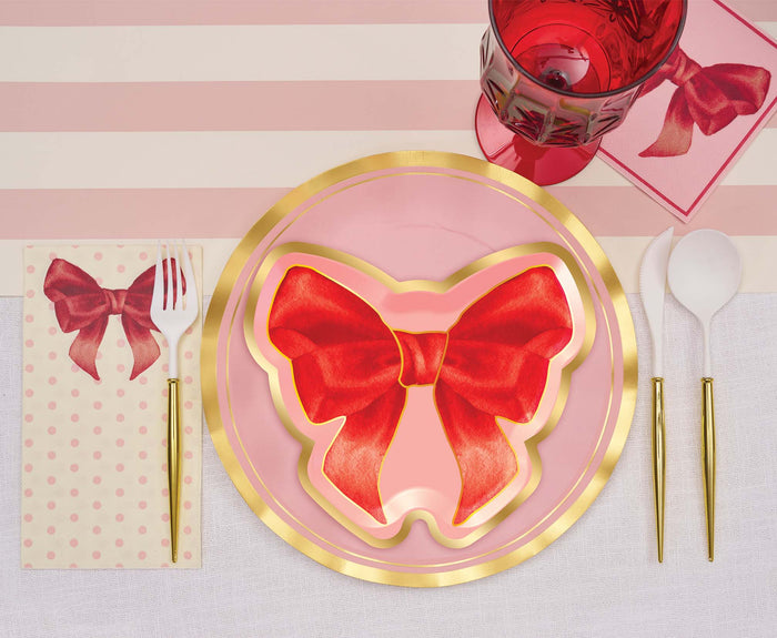 The Pink Bows Table Setting by Sophistiplate features a pink and gold theme with designer disposable red bow-shaped plates, gold utensils, a red glass, and matching napkin and card decorated with red bow illustrations.