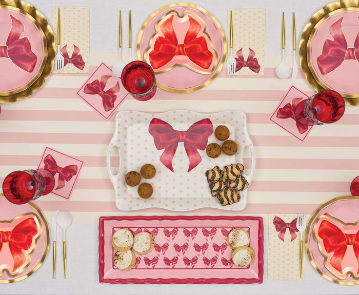 A festive Valentine’s Day table features Sophistiplate Pink Bows Assorted Die Cut Salad Plates (8pk), pink and gold accents, matching napkins, cups, trays with large red bows, cookies and desserts on a striped runner, and white utensils.