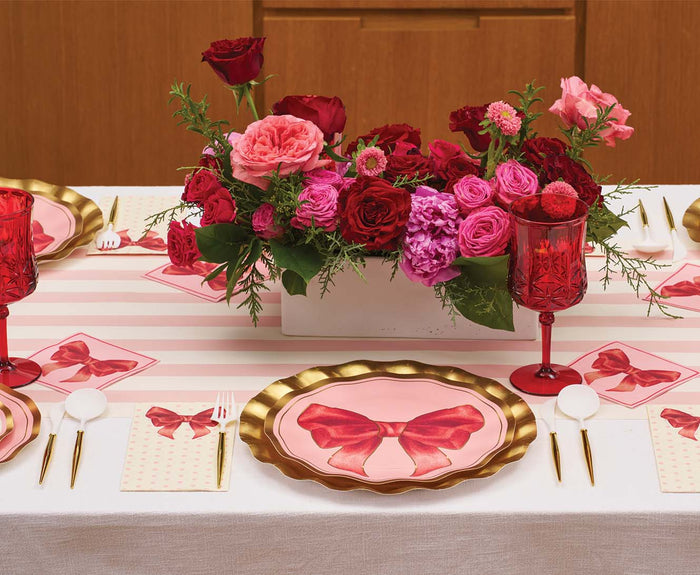 A table set for a party features Sophistiplate’s Pink Bows Dinner Plates/8pk from the Pink Bows Collection, adorned with gold edges and pink bows, along with red glasses, pink napkins, white cutlery, and a centerpiece of pink and red flowers. The birthday party tableware is complemented by a white tablecloth topped with a pink striped runner.