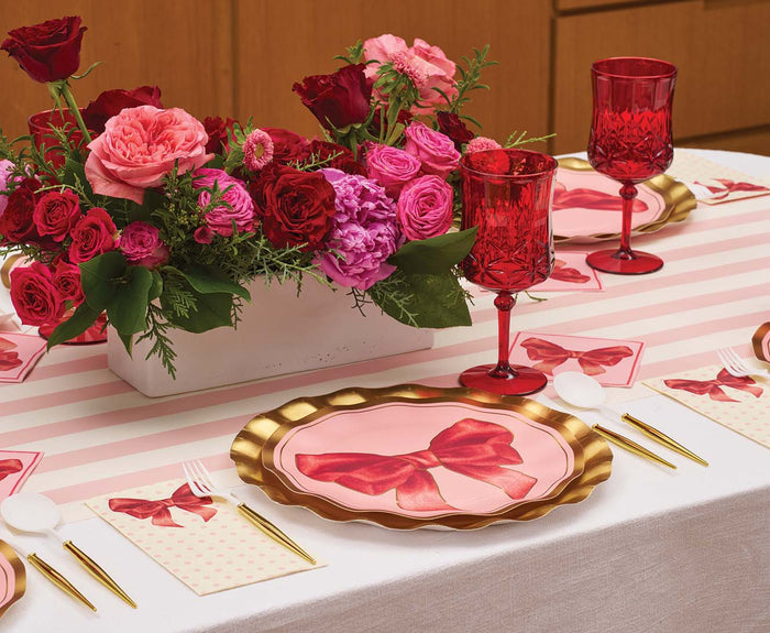 A table is set with Sophistiplate’s Pink Bows Dinner Plates (8pk), accented by red goblets, gold flatware, and a centerpiece of red and pink flowers. Featuring charming bow designs and stripes from the Pink Bows Collection, the decor creates an elegant, festive atmosphere—perfect for showcasing chic birthday party tableware or stylish disposable party plates.