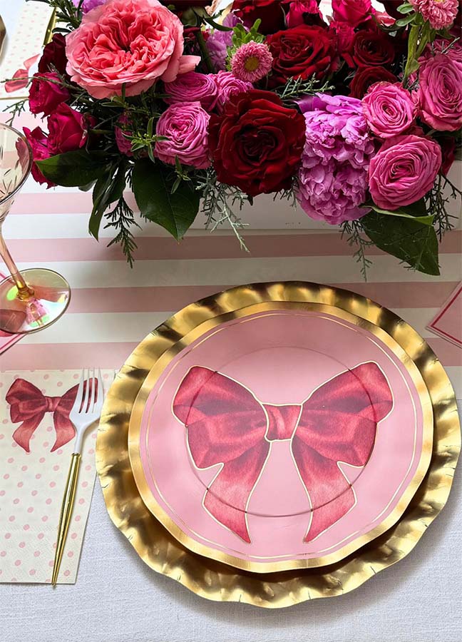 The Pink Bows Dinner Plates/8pk by Sophistiplate, featuring a charming red bow design from the Pink Bows Collection, are displayed on a gold charger beside a festive polka dot napkin and fork. Above, a bouquet of pink and red flowers adds an elegant touch, while a striped placemat enhances this delightful arrangement of disposable party plates—making them the perfect birthday party tableware for any celebration.