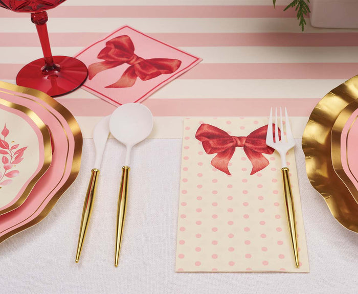 A festive table is set with pink and gold plates, gold-handled cutlery, a red wine glass, and Sophistiplate Pink Bows Guest Towels (20pk), featuring red bows and pink polka dots on a white and pink striped tablecloth.