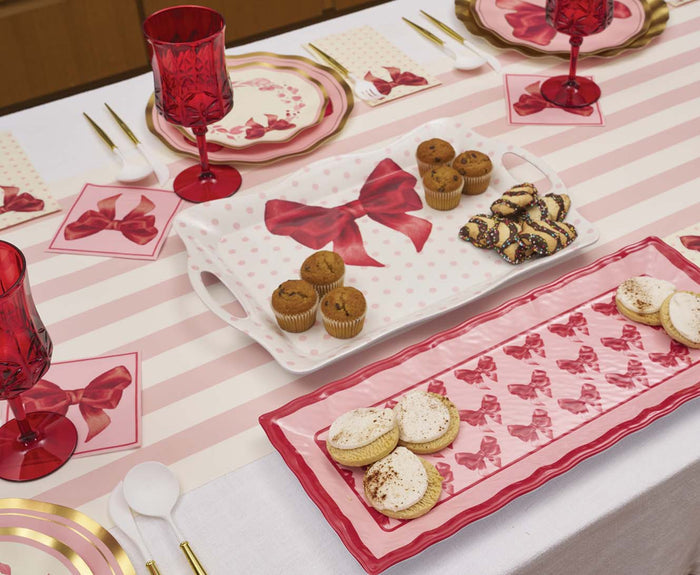 A festive table features a Sophistiplate Pink Bows Melamine Serving Tray, polka dot trays, red cups, gold flatware, and sweet treats arranged on a striped tablecloth.