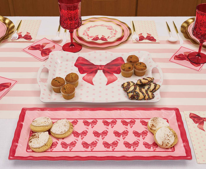 A Sophistiplate Pink Bows Melamine Serving Platter holds cookies and muffins, accompanied by neatly arranged plates, napkins, utensils, and red goblets on the table.