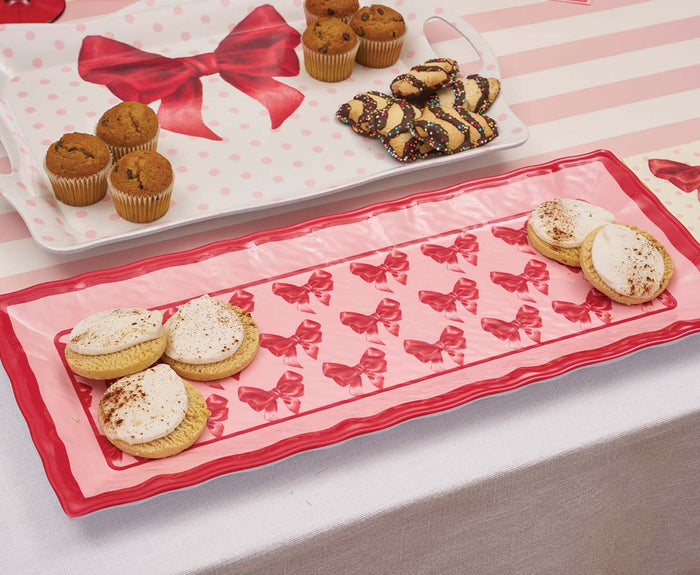 An assortment of baked treats, such as cream-topped cookies, sprinkle chocolate-dipped cookies, and muffins, is displayed on the Sophistiplate Pink Bows Melamine Serving Platter featuring red bows on a pink and white background.