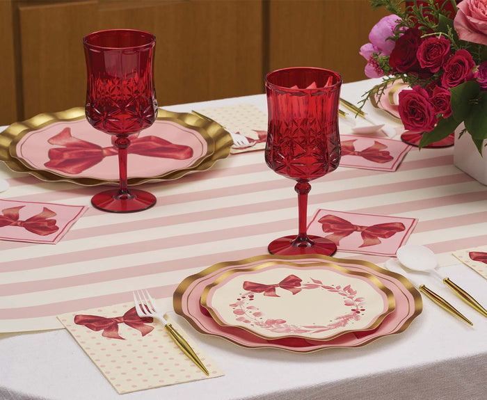Table set for two with red glasses, gold utensils, bow-themed napkins, and a bouquet of pink and red flowers on a striped tablecloth, featuring Sophistiplate Pink Bows Salad Plates (8-pack).