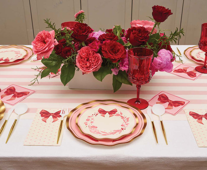A table set with Sophistiplate Pink Bows Salad Plates from the Pink Bows Collection, gold cutlery, red glasses, and pink napkins featuring bow designs. A centerpiece of red and pink roses is arranged in the middle on a pink-striped table runner.