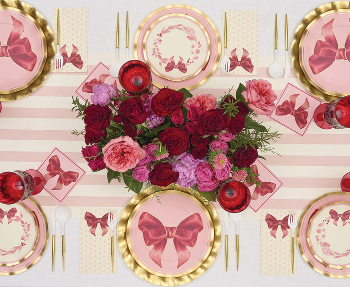 Table set with Sophistiplate Pink Bows Salad Plates from the Pink Bows Collection, featuring charming bow designs, paired with gold utensils, pink glasses, and a centerpiece of red and pink flowers on a pink striped table runner.