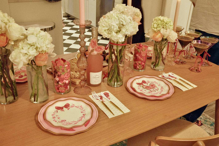 A wooden table set for a celebration features elegant floral arrangements, heart-shaped décor, a pink bottle, gold utensils, and cocktail glasses. The stylish and festive setup includes Sophistiplate Pink Bows Salad Plates (8pk), beautifully displayed on the table alongside other disposable party accents. A checkerboard floor in the background completes the sophisticated look.