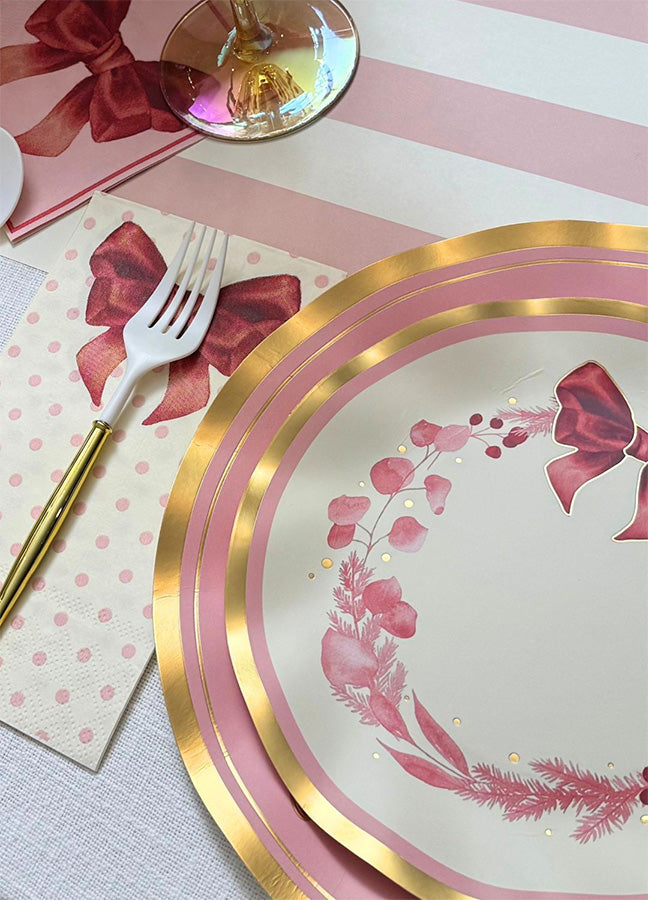 A close-up of a table setting from Sophistiplate’s Pink Bows Salad Plates (8-pack) showcases the brand’s pink and gold disposable party plates, a napkin adorned with a red bow, a gold fork, and a glass. The decor highlights festive floral elements, bows, and playful striped and dotted patterns.