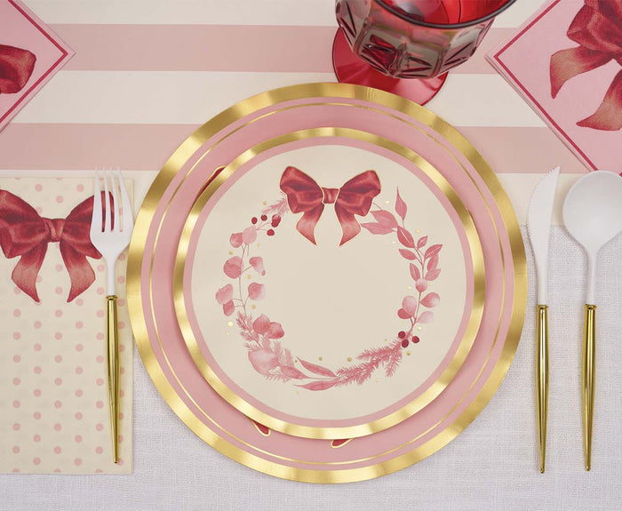 The Pink Bows Table Setting by Sophistiplate features a luxury paper plate with a bow and floral wreath design, white and gold utensils beside a bow-print napkin, and a pink glass on a striped tablecloth.
