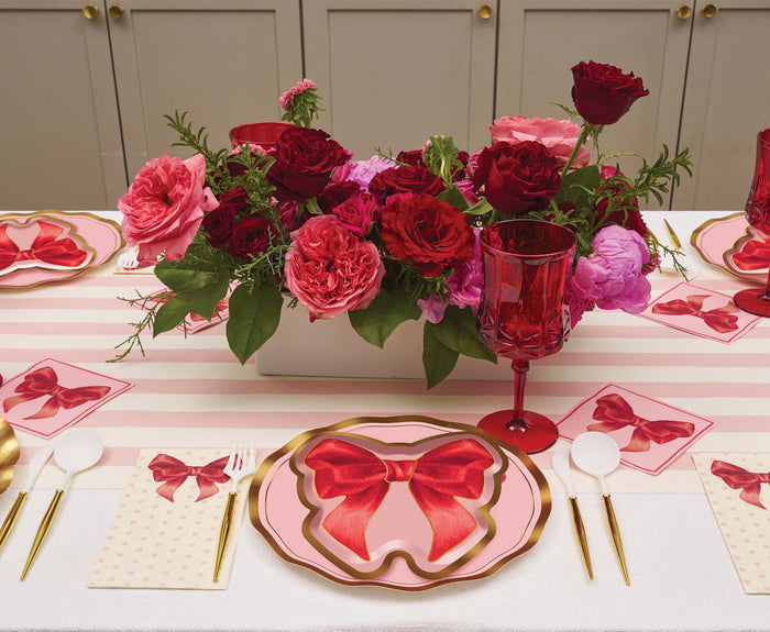 The Pink Bows Table Setting by Sophistiplate features designer disposable plates with pink and red bows, red glasses, white cutlery, pink napkins, and a red and pink floral centerpiece on a pink striped tablecloth.
