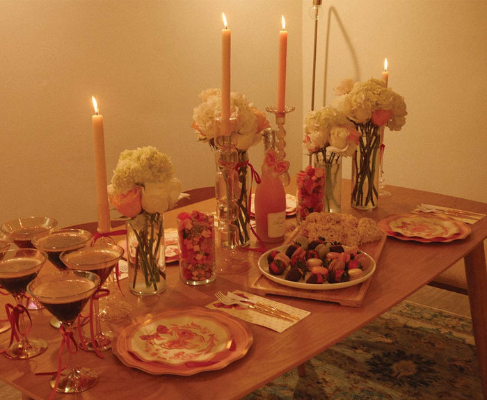 The Pink Bows Table Setting by Sophistiplate showcases a warmly lit wooden table adorned with floral arrangements, tall candles, chocolate-dipped strawberries, assorted pastries, and festive drinks for a romantic celebration.