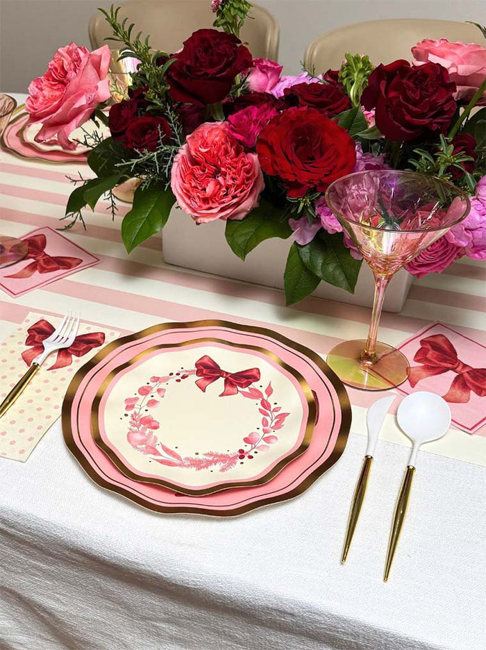 The Pink Bows Table Setting by Sophistiplate features pink and gold bow-accented luxury paper plates, white and gold cutlery, a pink stemmed glass, floral napkins, and a centerpiece of pink and red roses on a white cloth with pink striped runners.